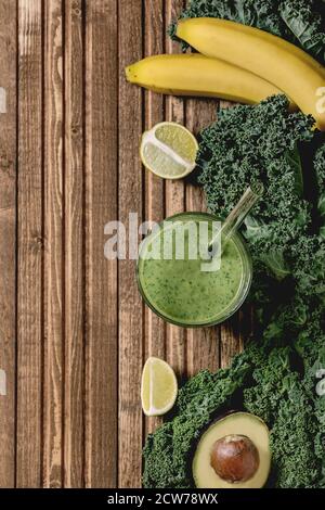 Verre de smoothie végétalienne saine et verte, paille de verre. Ingrédients ci-dessus. Kale, bananes, avocat, chaux, sur fond de planche en bois. Plat, espace Banque D'Images