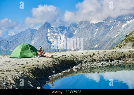 Mâle touriste assis dans une chaise près de la tente dans les montagnes alpines rocheuses près du lac avec de l'eau claire et fraîche et en utilisant un smartphone dans la chaude journée d'été ensoleillée. Concept de camping, voyage, nature, gadgets. Banque D'Images