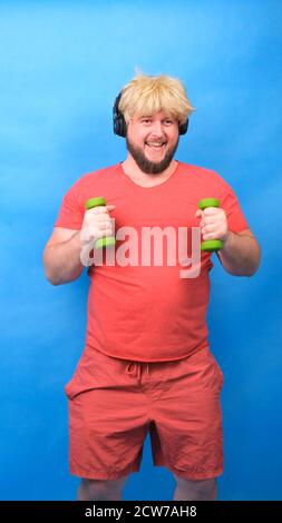 Drôle de chubby freak homme dans le casque et une perruque dans un t-shirt rose est engagé dans les haltères vertes et rires sur un fond bleu, vertical Banque D'Images