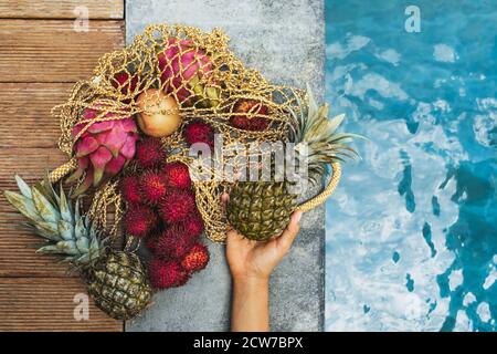 Femme tient main l'ananas de tas de fruits tropicaux frais dans un sac en tissu réutilisable sur le bord de la piscine. Nourriture en vitamines d'été. Aliments crus sains et régime végétarien. Aucun gaspillage. Banque D'Images