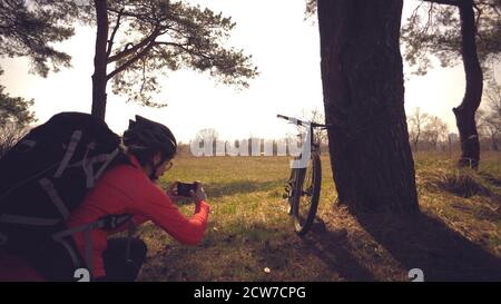 Jeune femme caucasienne athlète cycliste touristique fait photo au téléphone, photographies son vélo debout près de l'arbre sur le champ de la nature ensoleillé jour. Fille dans un casque Banque D'Images