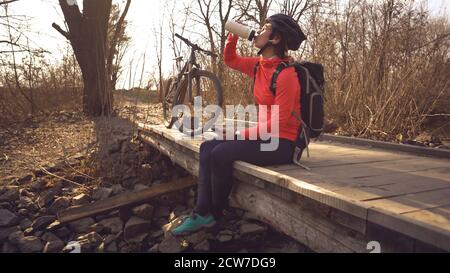 Happy Woman se trouve sur un pont en bois au-dessus de la rivière, écoute de la musique dans un casque avec les mains d'un smartphone et boit une bouteille d'eau blanche. Le cycliste écoute la chanson Banque D'Images