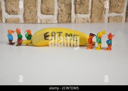 Banane. Fruit avec inscription, mangez-moi, avec des jouets en plastique miniature regardant le fruit, base blanche, fond de mur de brique, dans la photo conceptuelle, Brésil, Banque D'Images