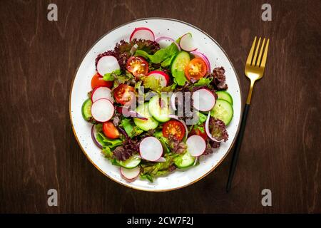 Salade de légumes sains de tomates cerises, tranches de concombre, feuilles de laitue verte et violette, oignons et huile d'olive sur une table en bois sombre Banque D'Images