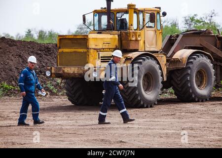 Deux ingénieurs ou travailleurs en bleu de travail et casques blancs sur grand fond jaune de tracteur. Gisement de pétrole de Zhaik-Munai, Kazakhstan. Banque D'Images