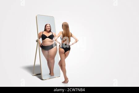 Jeune femme mince, en forme de femme regardant la grosse fille dans le miroir de réflexion sur fond blanc. Pensant qu'elle n'est pas assez sportive. Concept de mode de vie sain, forme physique, sport, nutrition et corps positif. Banque D'Images