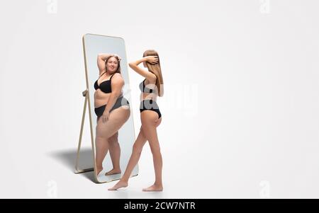 Jeune femme mince, en forme de femme regardant la grosse fille dans le miroir de réflexion sur fond blanc. Pensant qu'elle n'est pas assez sportive. Concept de mode de vie sain, forme physique, sport, nutrition et corps positif. Banque D'Images