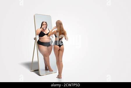 Jeune femme mince, en forme de femme regardant la grosse fille dans le miroir de réflexion sur fond blanc. Pensant qu'elle n'est pas assez sportive. Concept de mode de vie sain, forme physique, sport, nutrition et corps positif. Banque D'Images