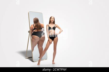 Jeune femme mince, en forme de femme regardant la grosse fille dans le miroir de réflexion sur fond blanc. Pensant qu'elle n'est pas assez sportive. Concept de mode de vie sain, forme physique, sport, nutrition et corps positif. Banque D'Images