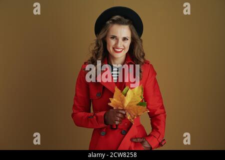 Bonjour octobre. Bonne femme tendance de 40 ans en manteau rouge et béret noir avec gants en cuir et feuilles d'érable jaune d'automne sur fond beige Banque D'Images