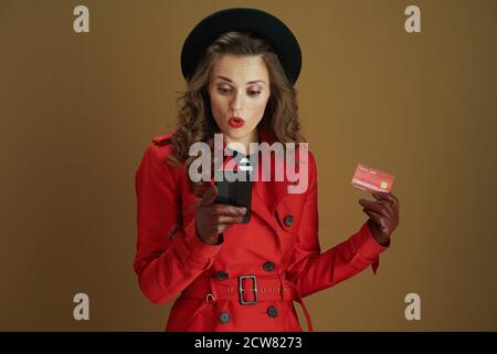 Bonjour octobre. Surprise élégante femme de 40 ans en manteau rouge et béret noir avec smartphone, carte de crédit et gants en cuir isolés sur le dos beige Banque D'Images
