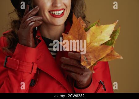 Bonjour automne. Gros plan sur une femme heureuse avec des gants en cuir et des feuilles d'érable jaune d'automne parlant sur un smartphone sur fond marron. Banque D'Images