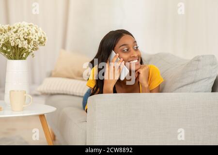 Femme noire gaie, couchée sur un canapé confortable avec un smartphone et bavardant avec son ami, à l'intérieur Banque D'Images