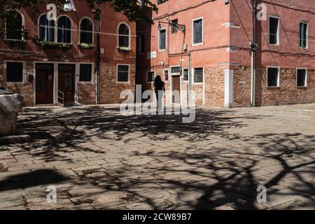 Lumière du soleil vive avec les ombres des arbres sur une petite place entourée de vieux bâtiments vénitiens de couleur rouge intense. Une jeune fille marche seule. Banque D'Images