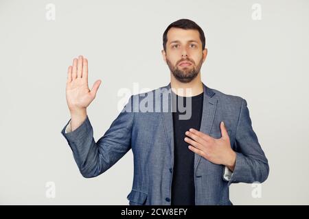 Jeune homme d'affaires avec une barbe dans une veste, jure, mettant sa main sur sa poitrine et paume ouverte, faisant un serment de loyauté. Portrait d'un homme sur fond gris. Banque D'Images