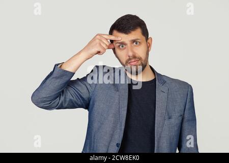 Jeune homme d'affaires avec une barbe dans une veste, pointant vers un bouton malheureux sur son front, une infection d'acné laid. Acné et problèmes de peau. Portrait d'un homme sur fond gris. Banque D'Images