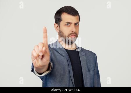 Jeune homme d'affaires avec une barbe dans une veste pointant son doigt vers le haut et expression de colère sans montrer de gestes. Portrait d'un homme sur fond gris. Banque D'Images