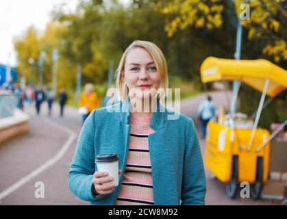 Femme blanche blonde souriante avec une tasse de café dans le parc d'automne. Portrait en plein air pendant l'été indien. Banque D'Images
