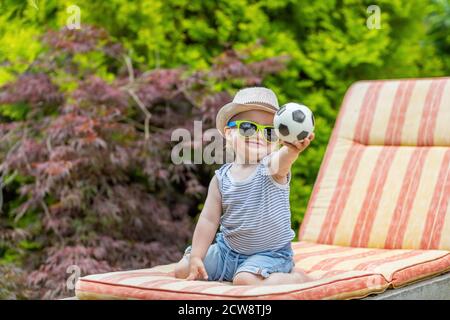 Le petit garçon heureux est assis sur la chaise de terrasse dans le jardin montrant le petit jouet ballon de football sur la caméra. Banque D'Images