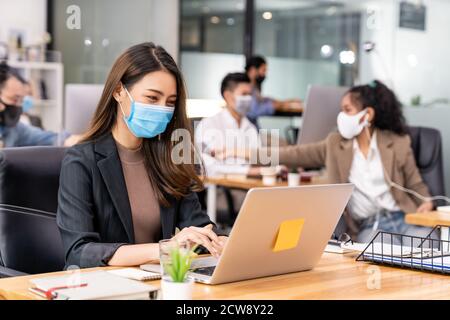 Portrait d'un employé de bureau asiatique femme d'affaires porter un masque de protection travailler dans un nouveau bureau normal avec une équipe interraciale en arrière-plan en tant que social dist Banque D'Images