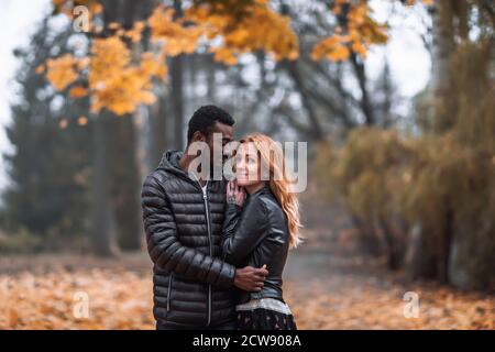 Heureux mignon couple interracial posant dans le parc d'automne flou, homme noir et femme rouge blanc Banque D'Images