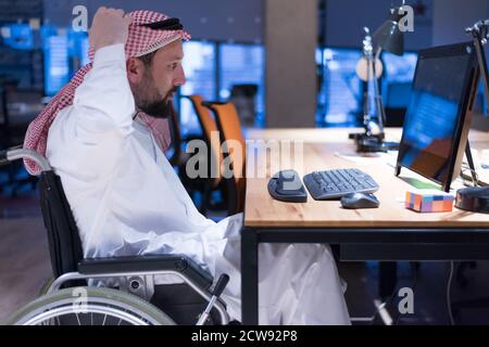 Homme d'affaires arabe handicapé dans la tradition costume travaillant dans le bureau. Homme d'affaires handicapé dans le fauteuil roulant travaille dans le bureau à l'ordinateur. Banque D'Images