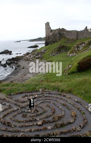 Femme de race blanche mature avec une coupe blonde de mulet en long manteau de cuir et voir à travers unbrella regardant autour des ruines du château et vieux village de pêcheurs à Ayrshire, en Écosse. A l'air remorquant et pensif.le jour est terne pluie menaçante qui s'inscrit dans ses pensées sombres, debout dans un labyrinthe de pierre regardant vers les ruines d'un vieux château Banque D'Images