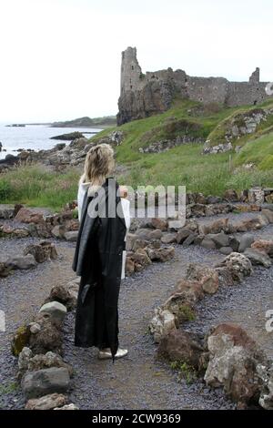 Femme de race blanche mature avec une coupe blonde de mulet en long manteau de cuir et voir à travers unbrella regardant autour des ruines du château et vieux village de pêcheurs à Ayrshire, en Écosse. A l'air remorquant et pensif.le jour est terne pluie menaçante qui s'inscrit dans ses pensées sombres, debout dans un labyrinthe de pierre regardant vers les ruines d'un vieux château Banque D'Images