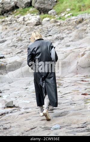 Femme de race blanche mature avec une coupe blonde de mulet en long manteau de cuir et voir à travers unbrella regardant autour des ruines du château et vieux village de pêcheurs à Ayrshire, en Écosse. Aspect remorquant et pensif.le jour est terne et menaçant de pluie qui s'intègre avec ses pensées sombres. Femme marchant sur la roche sur le rivage avec le château en ruines en arrière-plan Banque D'Images