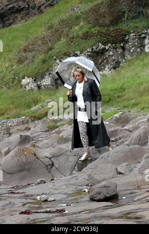 Femme de race blanche mature avec une coupe blonde de mulet en long manteau de cuir et voir à travers unbrella regardant autour des ruines du château et vieux village de pêcheurs à Ayrshire, en Écosse. Aspect remorquant et pensif.le jour est terne et menaçant de pluie qui s'intègre avec ses pensées sombres. Femme marchant sur la roche sur le rivage avec le château en ruines en arrière-plan Banque D'Images