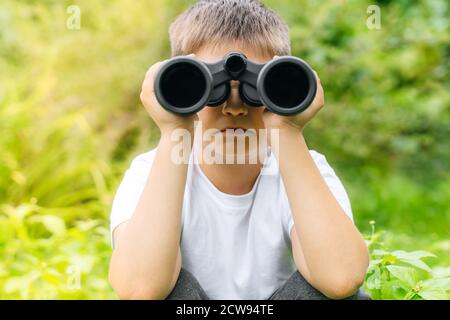 enfant garçon en t-shirt blanc regardant à travers les jumelles dans la nature vert paysage. Explorez et découvrez les concepts de la faune et de la flore. Aventure pour enfants, recherche Banque D'Images