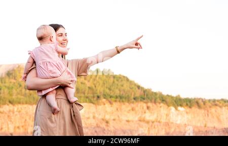 Une mère heureuse tient sa petite fille qui regarde loin et pointe vers l'horizon sur une carrière de coucher de soleil. Banque D'Images