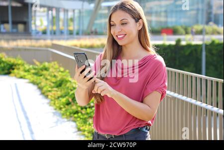 Jeune femme brésilienne caucasienne souriante, messages défilant sur l'application pour smartphone pour discuter dans la rue de la ville Banque D'Images