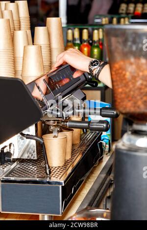 Un barista prépare le café dans une machine à café et le verse dans des tasses en papier à l'aide d'une cafetière et d'une pile de tasses en papier vides en arrière-plan. Banque D'Images