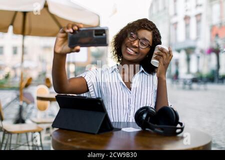 Charmante jeune femme africaine à la peau sombre en chemise rayée, faisant des selfie photo ou ayant appel vidéo, tout en étant assis dans un café extérieur de la ville, boire Banque D'Images