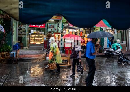 Un balai de rue dans un imperméable vert clair est occupé à nettoyer tandis que les navetteurs marchent avec leurs parasols dans le quartier chinois de Bangkok. Banque D'Images