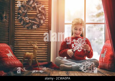 Joyeux Noël et joyeuses fêtes! Jolie petite fille assise près de la fenêtre et faisant des flocons de neige en papier. Chambre décorée. Enfant aime la chute de neige. Banque D'Images