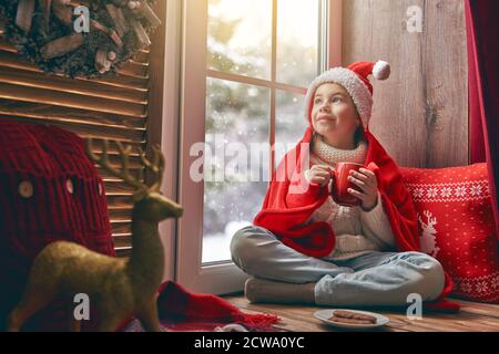 Joyeux Noël et joyeuses fêtes! Jolie petite fille assise près de la fenêtre avec une tasse de boisson chaude et regardant la forêt d'hiver. Chambre décorée. Banque D'Images