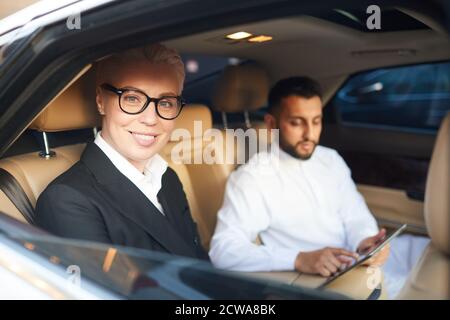 Femme d'affaires mature en lunettes souriant à l'appareil photo tout en étant assise la voiture avec son collègue en arrière-plan Banque D'Images