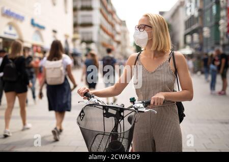 Femme marchant à vélo dans la rue piétonne de la ville portant un masque médical en public pour empêcher la propagation du virus corona. Nouvelle normale pendant Banque D'Images