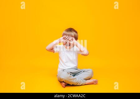 Le petit garçon pleure frotte ses yeux avec ses mains, un garçon dans un t-shirt blanc sur fond jaune Banque D'Images