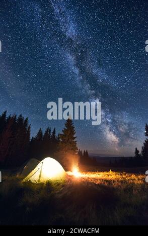 Feu de camp et tente illuminée sous un ciel étoilé lumineux sur lequel la voie lactée est clairement visible. Camping en soirée dans la vallée avec de grands pins. Ciel nocturne magique Banque D'Images