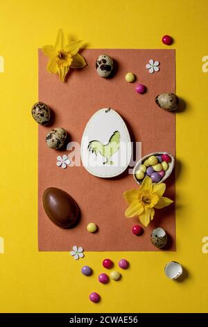 Carte de vœux de Pâques avec décorations en bois, bonbons au chocolat et œufs, jonquilles sur fond jaune orange vif. Couche plate, espace de copie. Banque D'Images