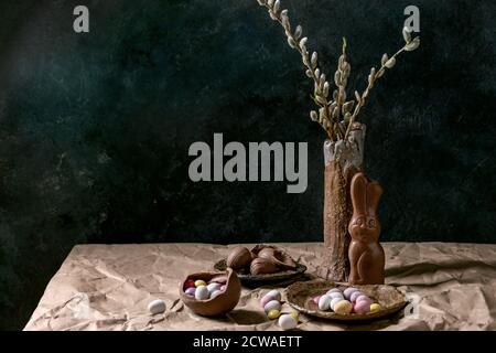 L'humeur de Pâques reste encore à vivre avec des branches de saule en fleur dans un vase en céramique, un lapin au chocolat traditionnel, des œufs et des bonbons sur la table avec du papier artisanal froissé. Banque D'Images