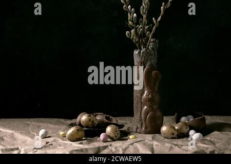 L'humeur de Pâques reste encore à vivre avec des branches de saule en fleur dans un vase en céramique, un lapin au chocolat traditionnel, des œufs et des bonbons sur la table avec du papier artisanal froissé. Banque D'Images