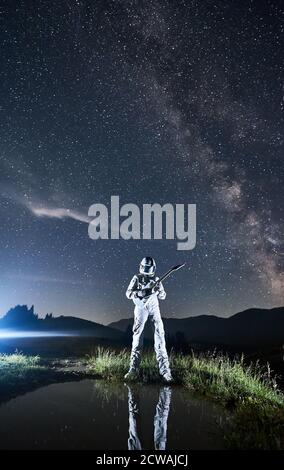 Spaceman dans un costume d'espace et un casque jouant de la guitare sous le ciel bleu de nuit avec la voie lactée. Guitariste cosmonaute avec instrument de musique réfléchissant dans un petit lac. Concept de musique, de cosmonautics et de galaxie. Banque D'Images