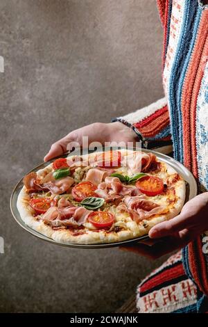 Pizza maison fraîche avec jambon prosciutto, fromage, tomates, basilic sur plaque dans les mains des femmes. Cuisson à domicile ou livraison de fast-food. Banque D'Images