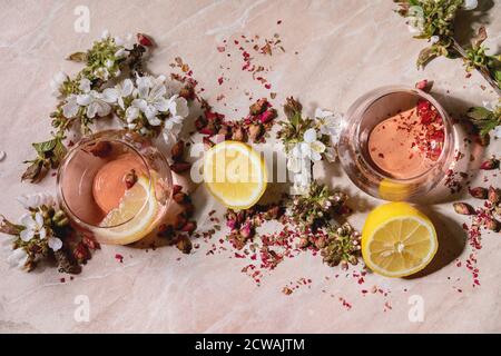 Verres à cocktail de champagne rose, de cidre ou de limonade avec boutons de rose secs, citron et menthe. Des branches de cerisier fleurient au-dessus. Fond en marbre rose. Banque D'Images