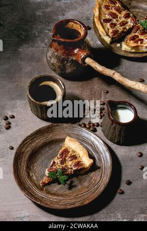 Morceau de maison de cuisson de tarte de pacan américain classique au caramel sur plaque de céramique, tasse de café, cezve, verseuse de crème sur fond de texture grise. Banque D'Images