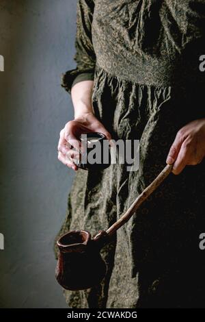 Femme en robe sombre tenir dans les mains artisanat céramique pot de café cezve avec une longue poignée en bois et un pot de lait pour faire du café turc. Banque D'Images